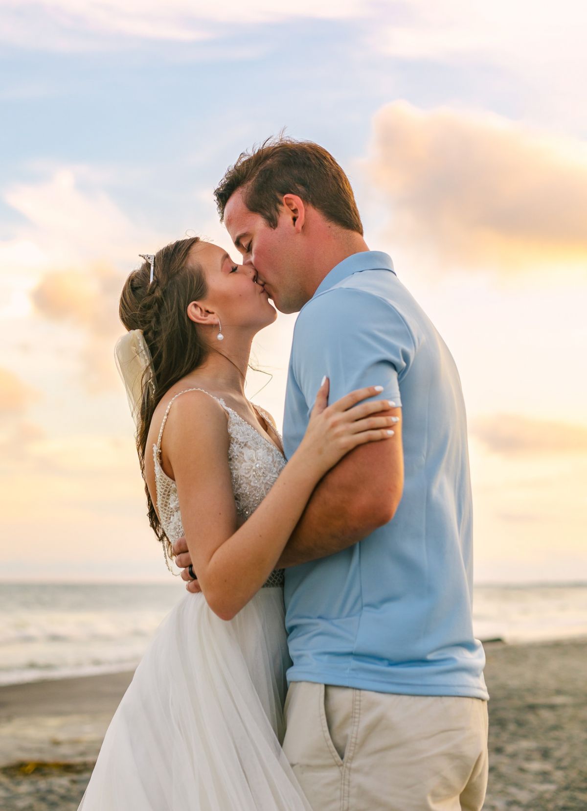 Couple kisses after wedding on the beach with pink clouds in background, Folly Beach