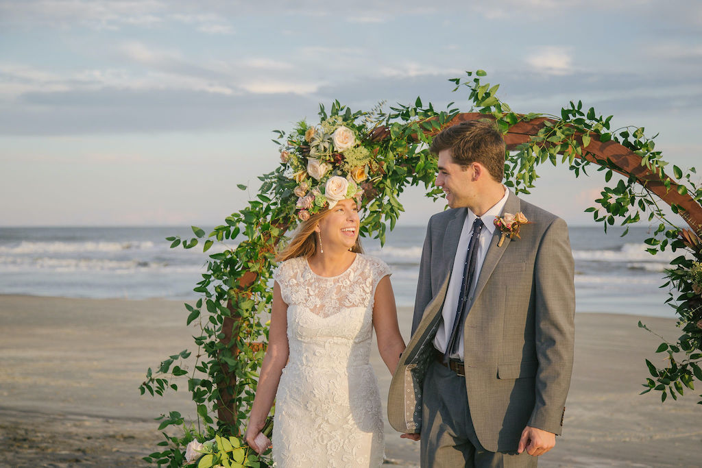 Folly Beach wedding with flower arbor on the beach near Charleston SC. Couple is looking at each other, one wears a flower crown